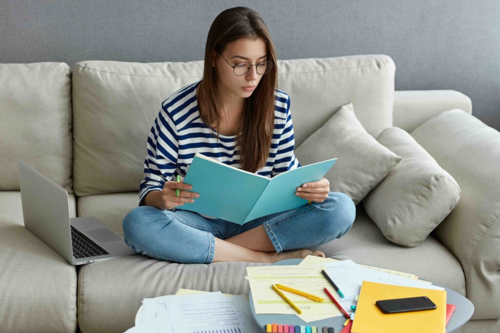Young freelancer does ditance job at home, studies literature, learns foreign language in internet, writes down unknown words, sits crossed legs on couch, spends time at home, connected to wifi