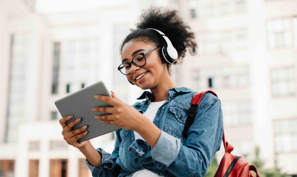 E-Learning. Happy Black Woman Student Using Tablet Computer Wearing Headphones Standing Near Modern University Building Outside, Browsing Internet Outdoors. Modern Online Education, Copy Space