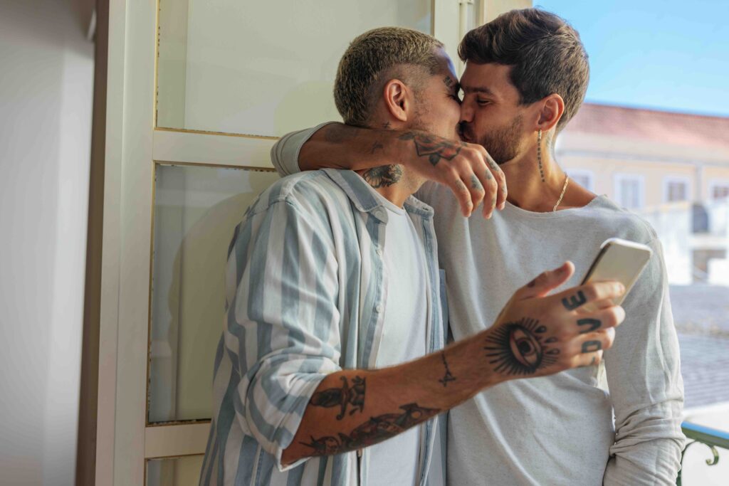 Happy tattooed gay couple kissing and taking a photo with a smart phone while standing on a balcony