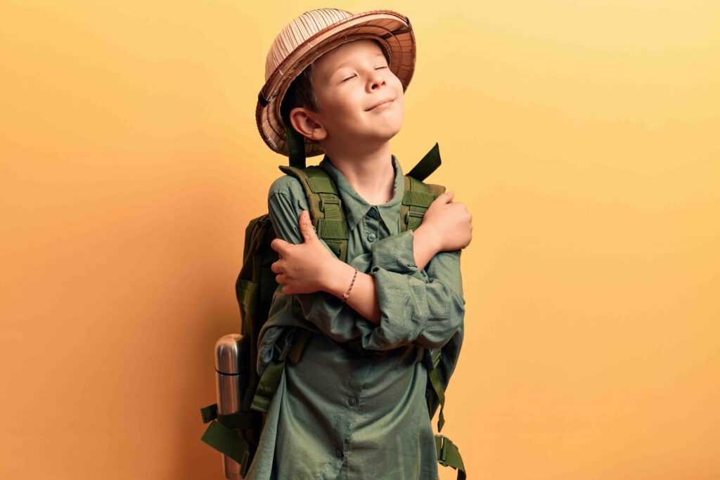 Cute blond kid wearing explorer hat and backpack hugging oneself happy and positive, smiling confident. self love and self care