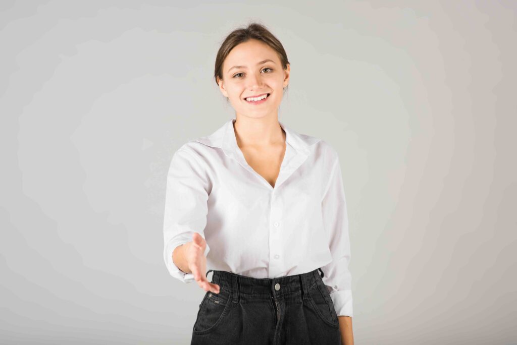 A young Caucasian brunette businesswoman in a white shirt reaches her hand for a handshake isolated on a gray studio background.