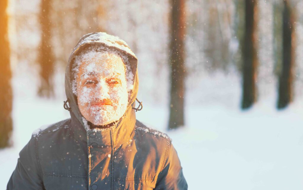 Fun portrait of an young frozen man. Jogging in a blizzard in the woods. Face covered with snow and frost. Closeup portrait of happy young guy smiles in cold weather in the winter forest at sunset