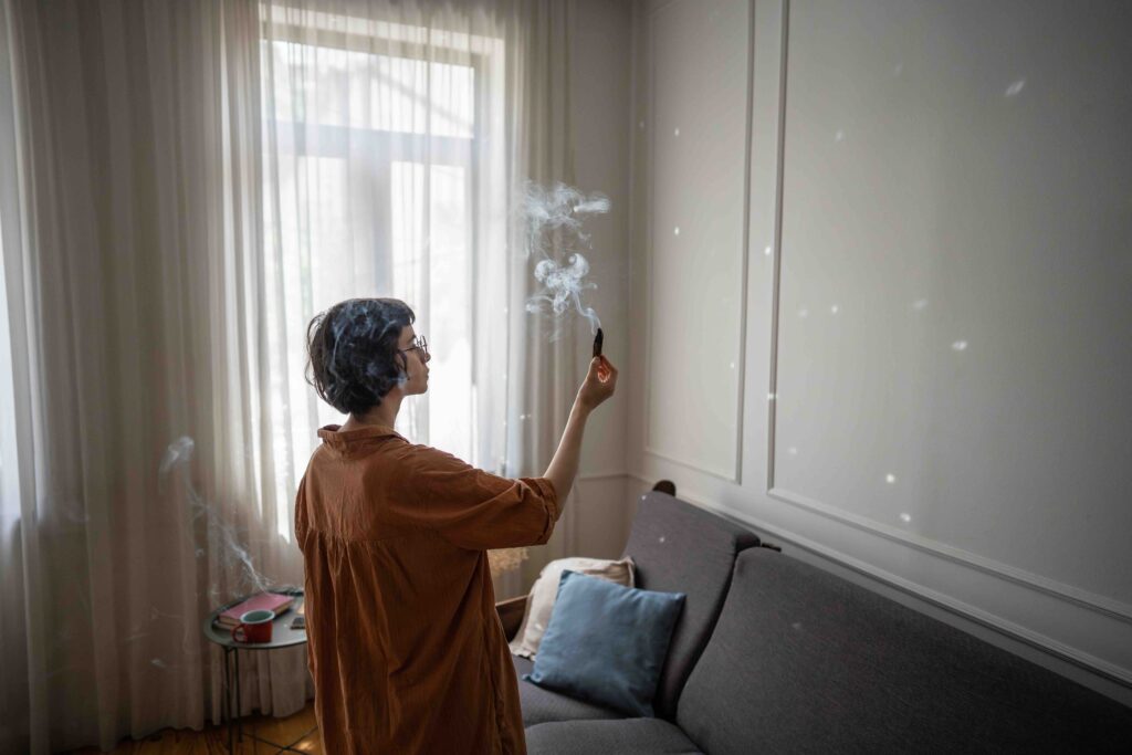 Calm woman lighting palo santo stick, filling home with aromatic smoke cozy atmosphere for meditation or work. Sacred ritual, natural mindfulness practice, space purification, emotional balance