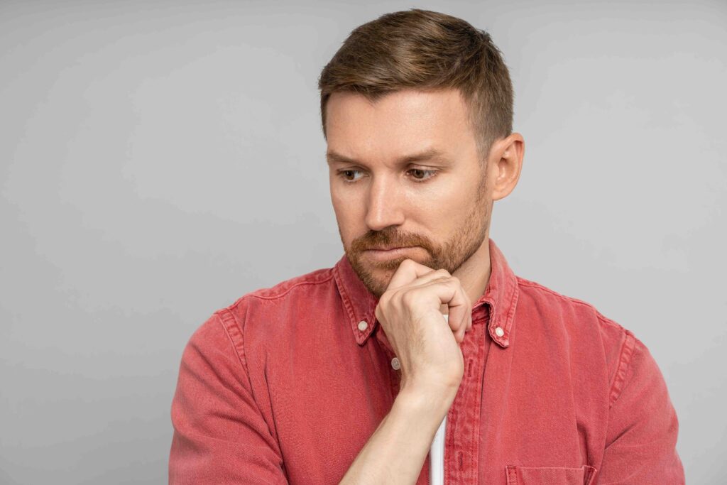 Sad pensive thoughtful man touching chin looking down isolated on gray background. Bearded serious middle aged caucasian blond guy wistfully thinking. Placard, poster. Sincere human emotions concept.
