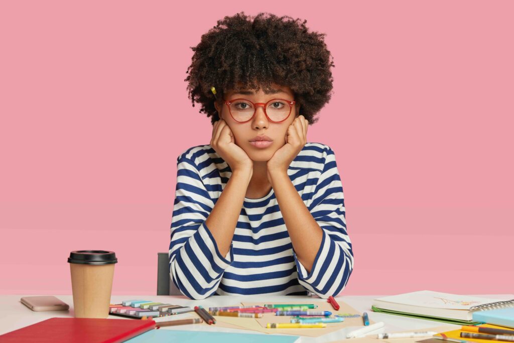 Sad dejected female architect feels unhappy as has no inspiration for creating masterpiece, sits at desktop, surrounded with crayons, notebook and disposable cup of coffee, holds chin isolated on pink