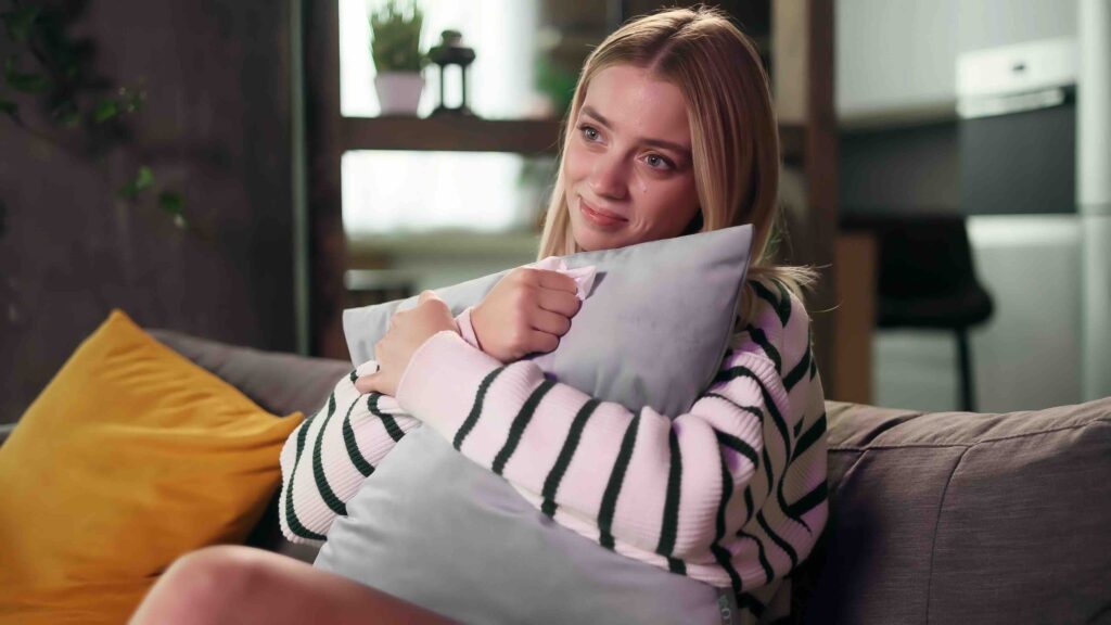 Portrait if sensitive young attractive woman sits on sofa at home alone hugging a pillow and watch drama film Beautiful blond girl wipes away tears after watching film
